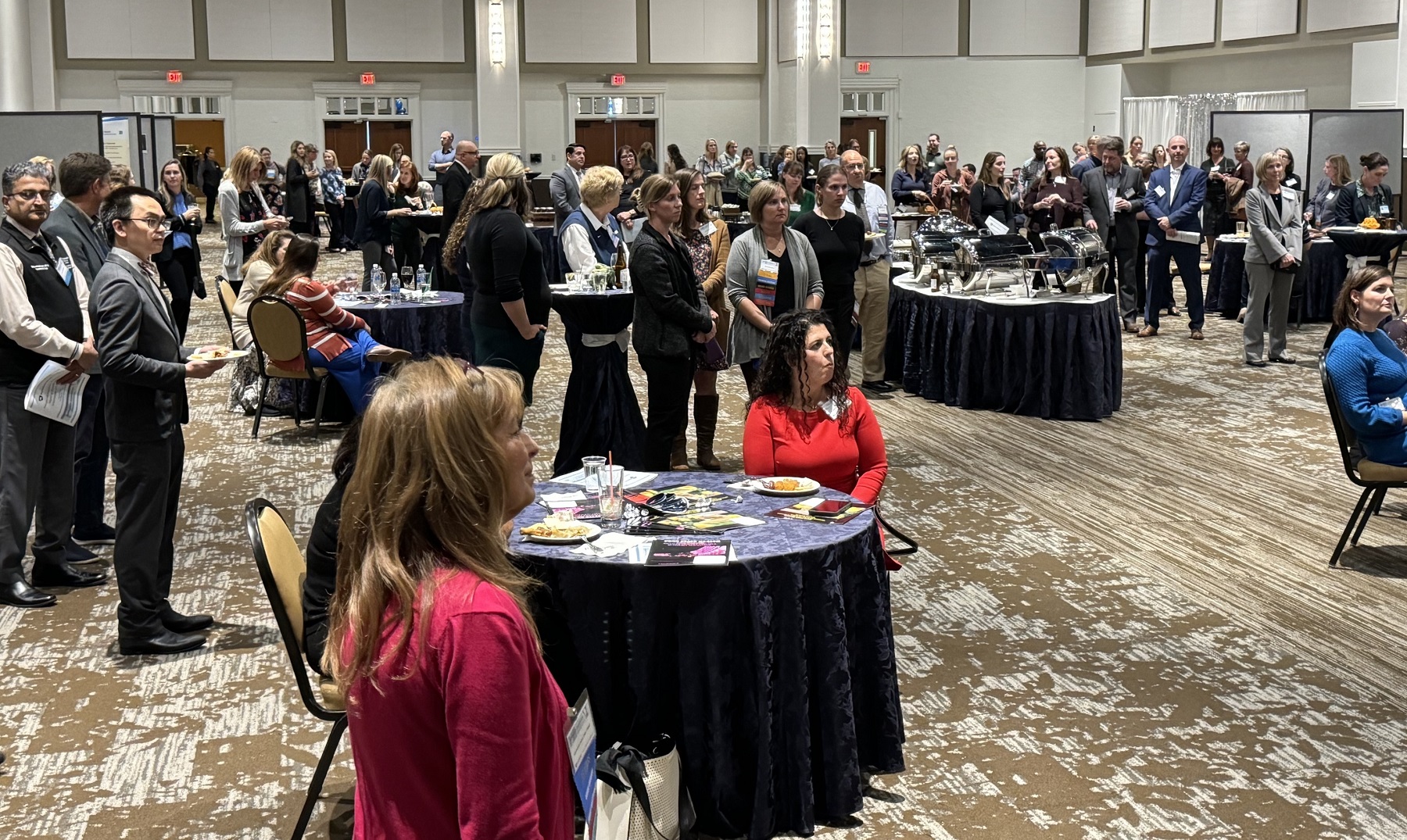 A large audience sits at round tables in a ballroom. They look to the right at the award presentation speaker. 