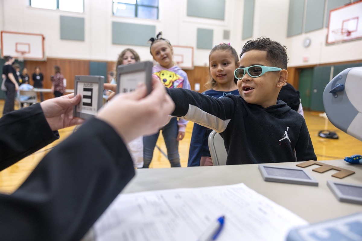 A little boy in glasses points at an image held by an adult. A line of children stand behind him.