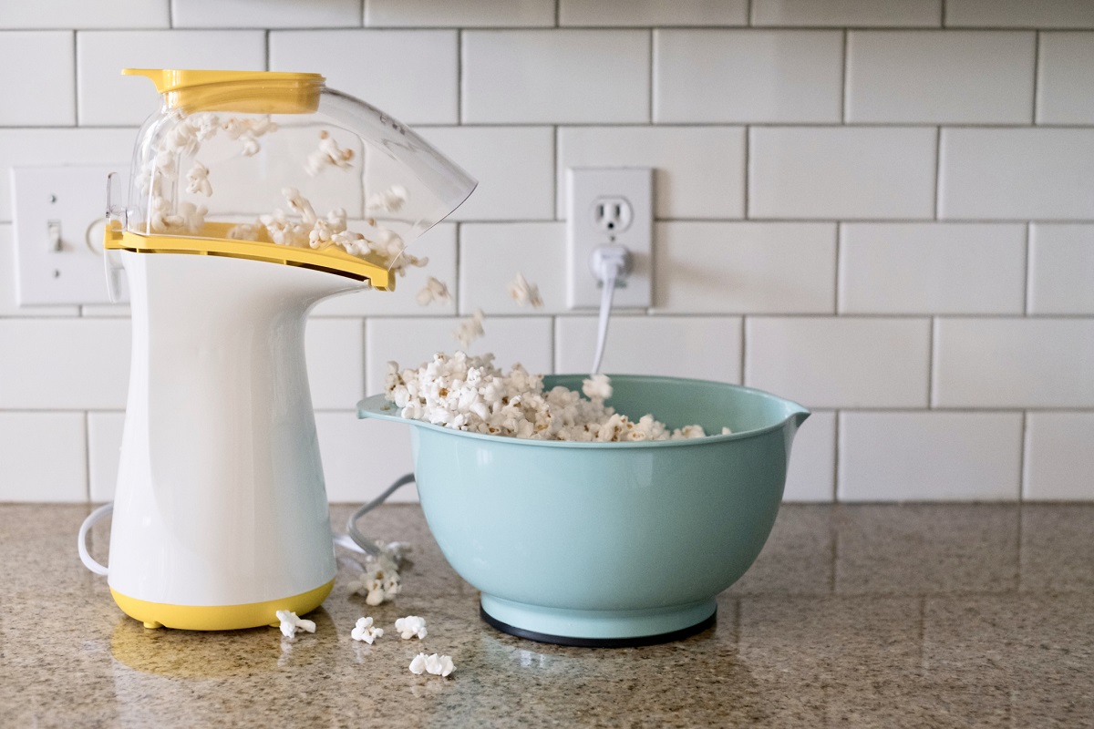 Popcorn machine popping popcorn into a aqua colored bowl on the countertop of a residential kitchen. The popcorn is overflowing onto the counter.