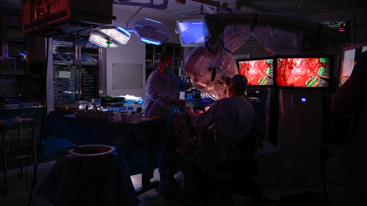 Interior of a darkened operating theater. Two people in scrubs stand before close-up video images of a brain.