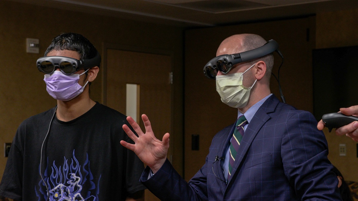 A boy in a T-shirt stands next to a man in a suit. Both wear goggles. The man is gesturing.