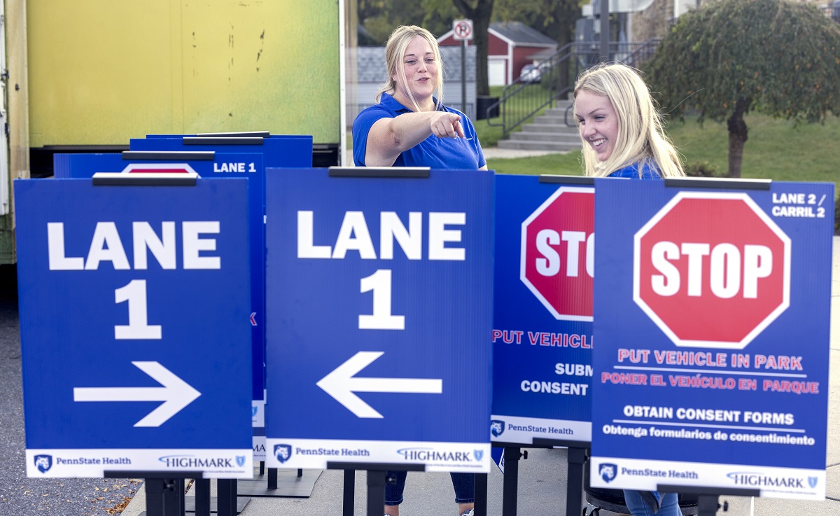 Two women stand amid signs that say Lane 1 with arrows. Others include stop signs. Both women smile and one points.