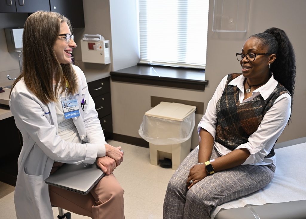 Dr. Allison Barrett smiles at patient Trina Love as they sit in Barrett’s office. Barrett is wearing a white coat and glasses. Love is wearing slacks with a blouse and vest.