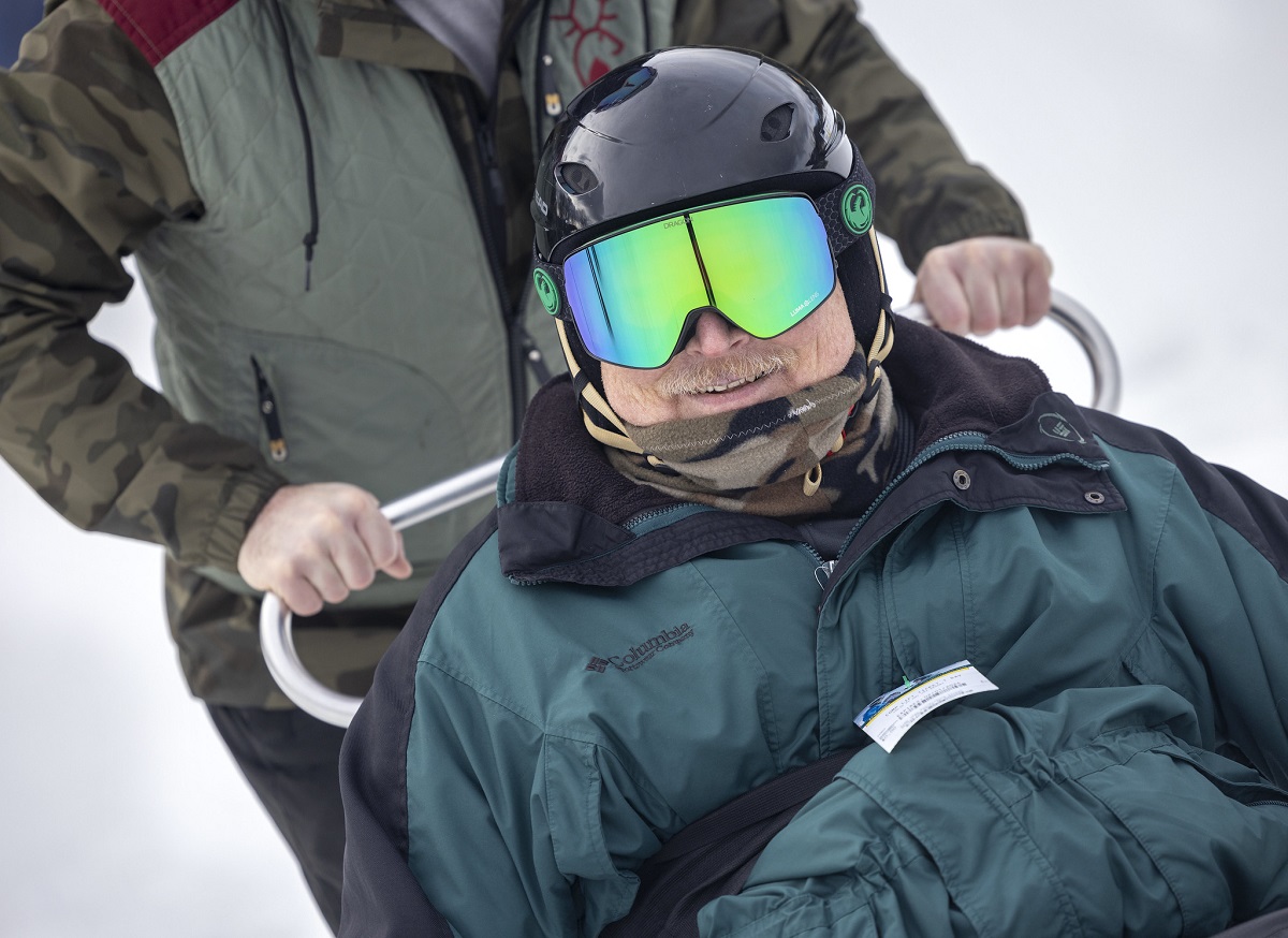 Close up of a man in ski goggles and a helmet. Beneath his mustache, he\'s smiling. Behind him, a pair of hands grip the handle of his sit ski.