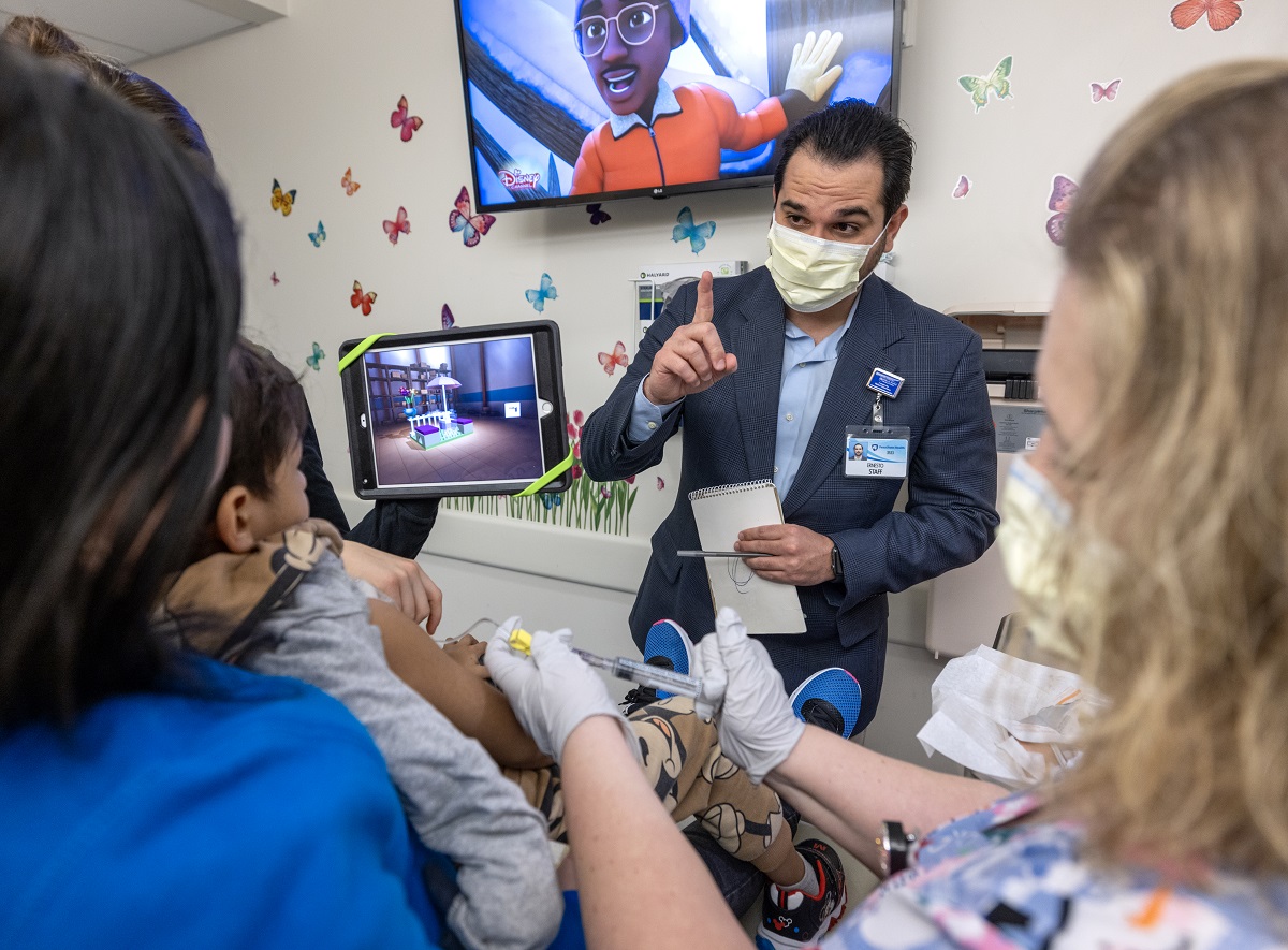 A man in a surgical mask and a sport jacket holds up an index finger while talking to three women in a hospital room.