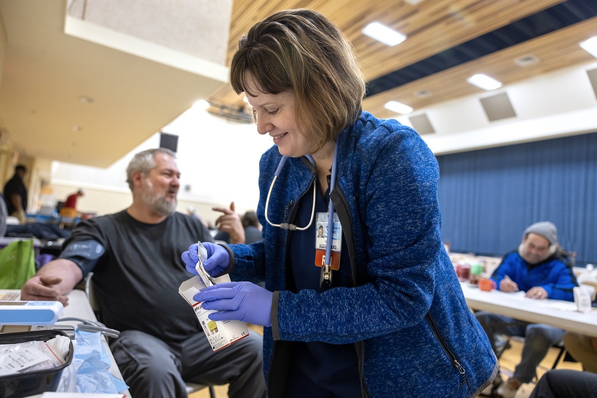 A woman wears rubber gloves and handles a box of medicine. A man wrests his arm, which is wrapped with a blood pressure cuff, on a table between them. Another man sits off to the side at another table, out of focus.