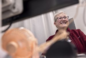 An overhead perspective of a cheerful medical staff member in glasses and a red jacket, with a nametag, standing in a clinic room, smiling at a patient out of view