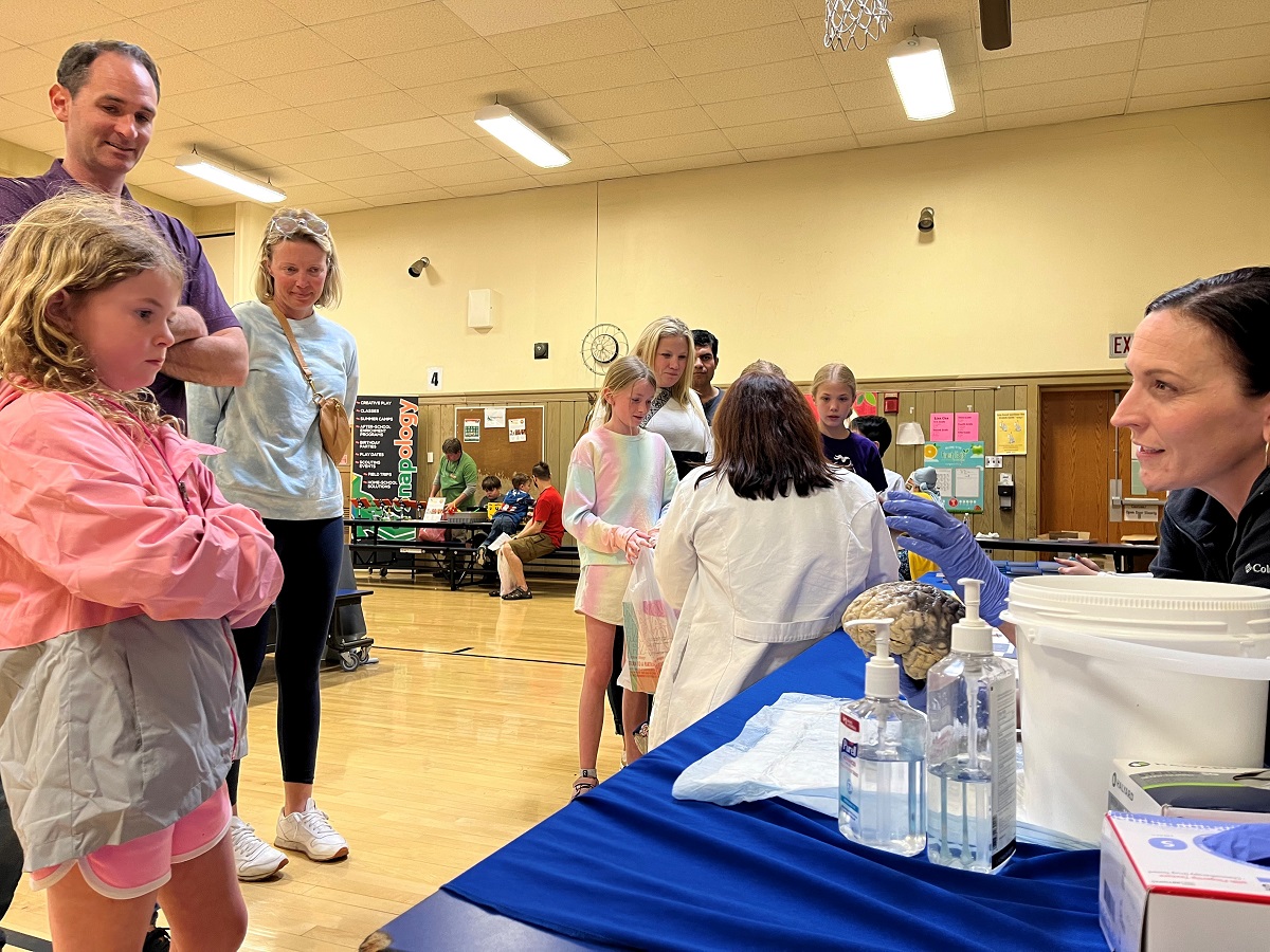 A woman in rubber gloves seated at a table shows a disembodied human brain to a child.