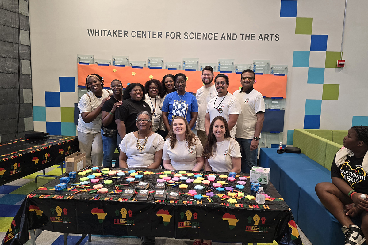 Twelve volunteers from Penn State Health smile and gather behind an activity table during the Juneteenth celebration at the Whitaker Center.